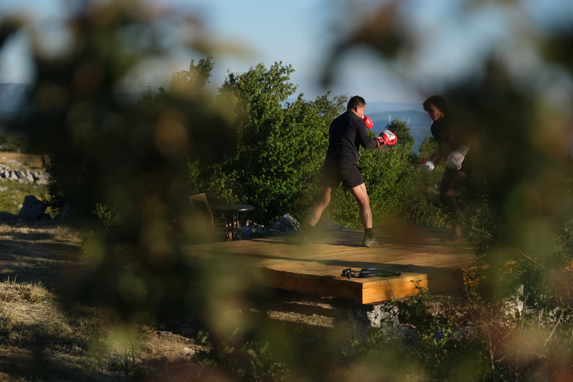 Two people sparring on the wooden deck at golden hour, the mountain ridge behind them